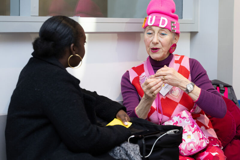 Two woman talk. One is wearing a black jacket the other is wearing a pink beanie and all pink/purple clothing