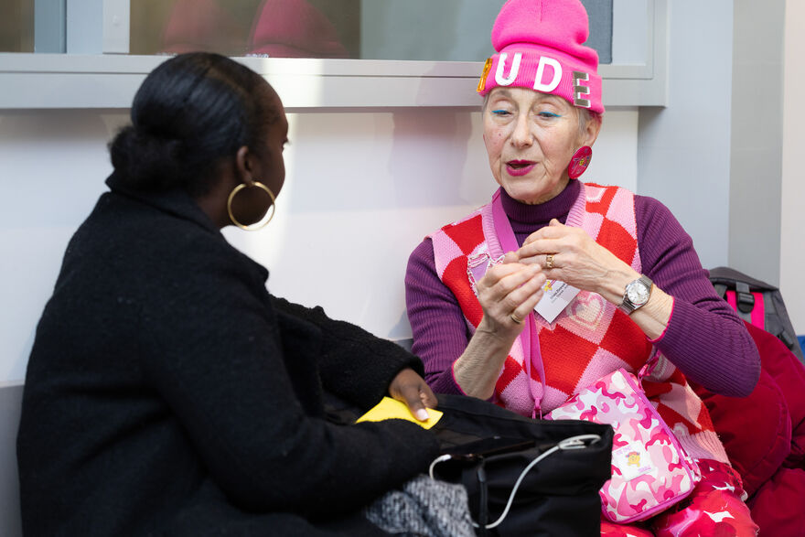 Two woman talk. One is wearing a black jacket the other is wearing a pink beanie and all pink/purple clothing