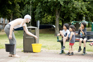 A white man with a yellow bucket perfoms in a park for three small children