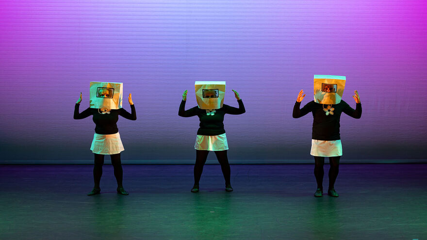 Three people on stage wearing white skirts, black tops, gold shape necklaces and gold boxes on their heads.