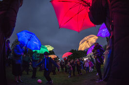 Lots of people with glowing colourful umbrellas outside at night time, a boy playing with a football amongst them