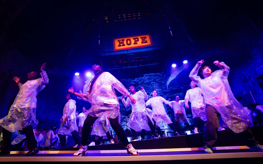 A group of young people dancing on stage. They are each wearing white t-shirts, black jeans, and a transparent poncho. There is a sing above them saying 'Hope'. 