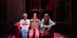 Three people are sat on a red sofa on a stage, surrounded by red and pink confetti. There is one man on the left-hand side in jeans and a t-shirt, he has blonde hair in a ponytail and green paint in war-like markings across his face. The other two are women – one on the right-hand side of the sofa is wearing a green hoodie and combat boots, and the one in the centre is wearing orange joggers and is holding a cardboard box.  Box Office