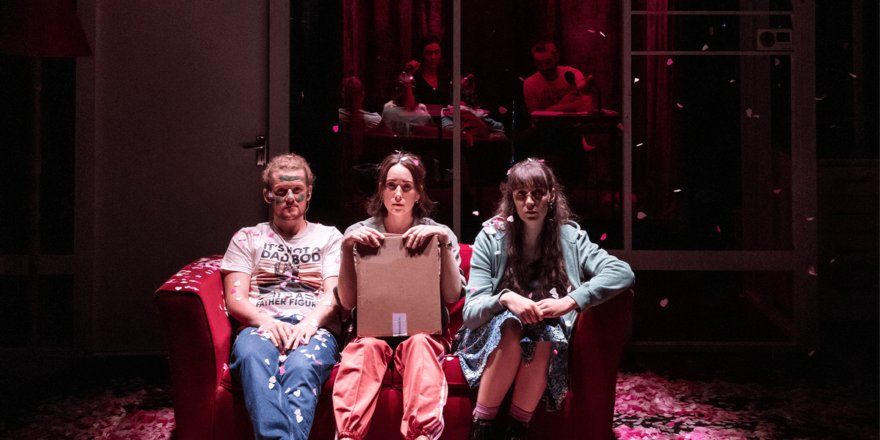 Three people are sat on a red sofa on a stage, surrounded by red and pink confetti. There is one man on the left-hand side in jeans and a t-shirt, he has blonde hair in a ponytail and green paint in war-like markings across his face. The other two are women – one on the right-hand side of the sofa is wearing a green hoodie and combat boots, and the one in the centre is wearing orange joggers and is holding a cardboard box.  Box Office