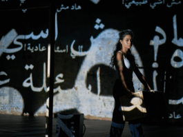 A woman stands holding a basket with Arabic script projected onto her.