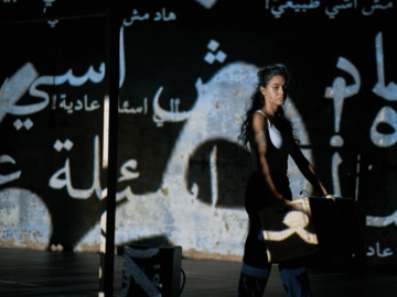 A woman stands holding a basket with Arabic script projected onto her.