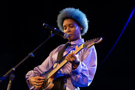 Person in purple jacket with blue afro playing guitar on stage 