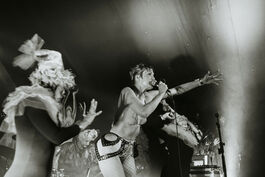 Black and white photo of person on stage, singing into microphone, wearing bikini top and fishnet tights