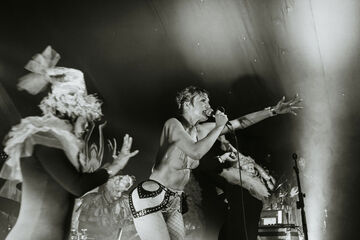 Black and white photo of person on stage, singing into microphone, wearing bikini top and fishnet tights
