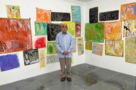 man stands infront of a collection of brightly coloured paintings hanging on the wall.