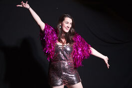 Caucasian woman in sparkly black jumpsuit and pink fringed shawl with arms stretched out, against black background