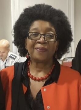 Photographic portrait of Dr Velma Mclymont, who is smiling and wearing an orange and black blouse/jacket, and orange beads.