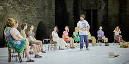 A group of people sitting in a semi-circle on a stage. The group is mixed gender and a mixed age range, wearing casual clothes. The group who are sitting down are audience participants in the show. There is also a male presenting person standing on the stage. The standing figure is Tiago, the artist who created the show and invited the audience participants on stage.