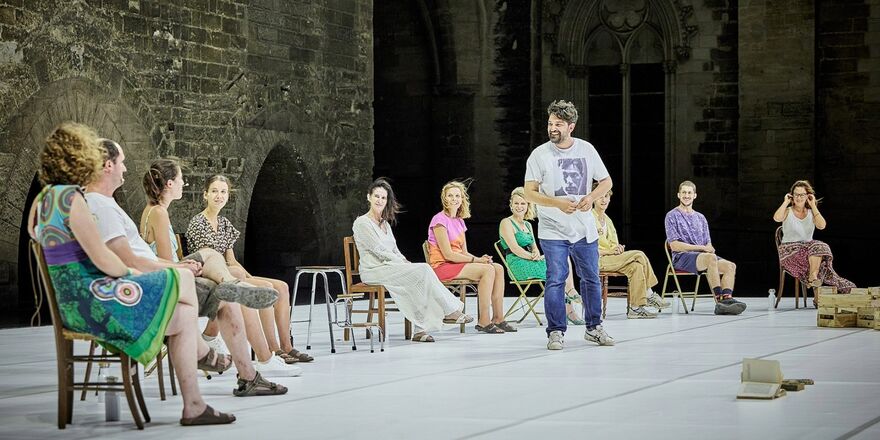 A group of people sitting in a semi-circle on a stage. The group is mixed gender and a mixed age range, wearing casual clothes. The group who are sitting down are audience participants in the show. There is also a male presenting person standing on the stage. The standing figure is Tiago, the artist who created the show and invited the audience participants on stage.