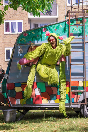a performer dressed in a fluffy green dragon suit and red sunglasses climbs out of the window of a colourful caravan