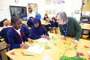 White woman leaning over classroom table, while two black young people do crafts, more children at tables in background