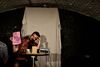 A man in a hawaiin shirt sits on a chair inside an arch. He is closing his eye and holding a pink piece of paper with a drawing on and iand the word 'arrrgghh'.