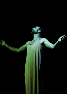Niall performing in a dress under a green light, with arms outstretched, looking up