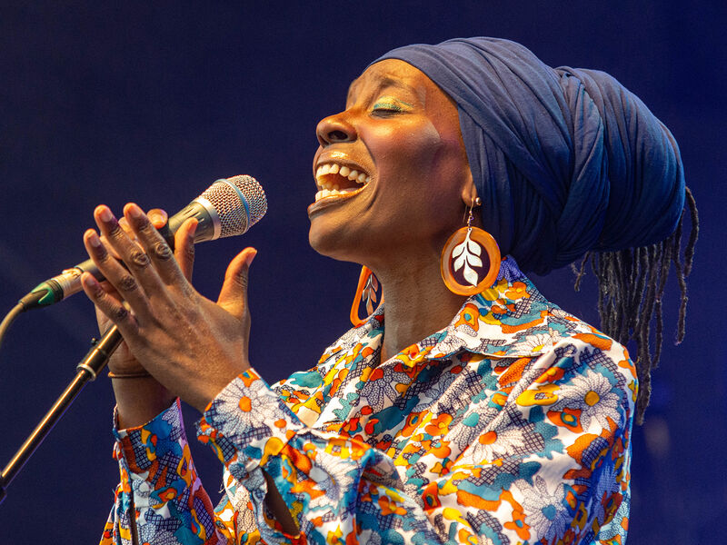 Publicity image of Singer, Melissa James, holding a microphone during a stage performance, wearing a colourful shirt, leaf earrings and blue headscarf