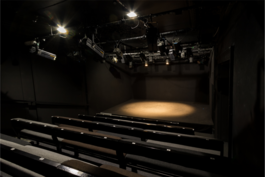 A spotlight on an empty black box stage, bench-style seating in the foreground