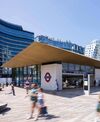 Modern underground station entrance with a large, flat roof and glass facade. People walk by on a sunny day. Nearby, curvy, high-rise buildings.