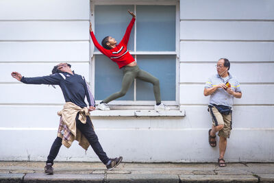 Three people posing outside a building. One has his arms and legs out and his head back while taking a step, another has stretched to fill the space of a square window and another is leaning against the wall, watching the others.