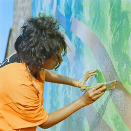 A woomna with black hair and an orange t-shirt paints a mural on a wall