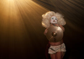 Midgitte Bardot, a person with dwarfism, performs on stage. They wear bold white makeup with blue eyeshadow covered mostly by thin white sunglasses, a glamorous blonde wig, red fish net tights and silky white shorts. They hold a cymbal in front of their chest which reflects golden light, with rays of more golden light flooding from the top left. Photo by Holly Revell. 
