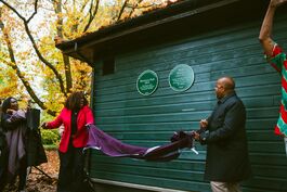 Black man and woman pull down purple covering from green plaque on side of outdoor building with trees behind them