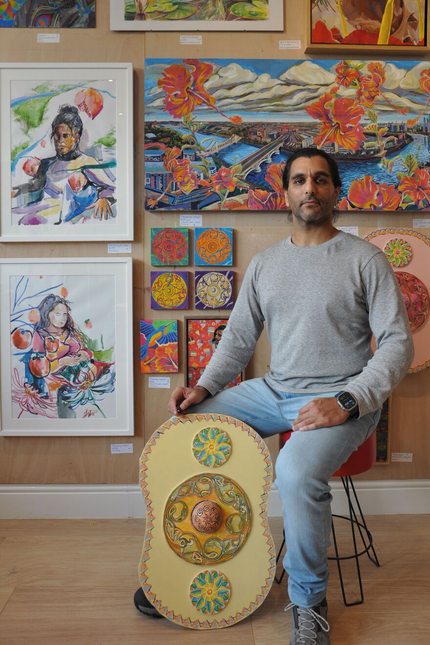 man sitting down in front of paintings on wall with a shield shaped sculpture resting against his leg