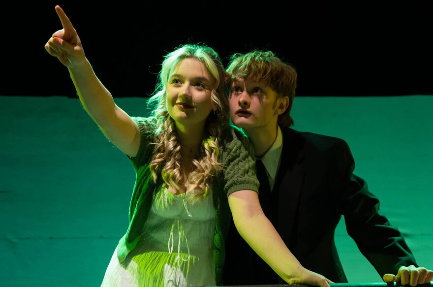 Two young people stand close to each other. One young person points to the left hand corner and the other looks where the point is pointing. 