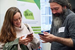 woman and man look at their phones laughing with a Re:Create pull-up banner in the backgrounf