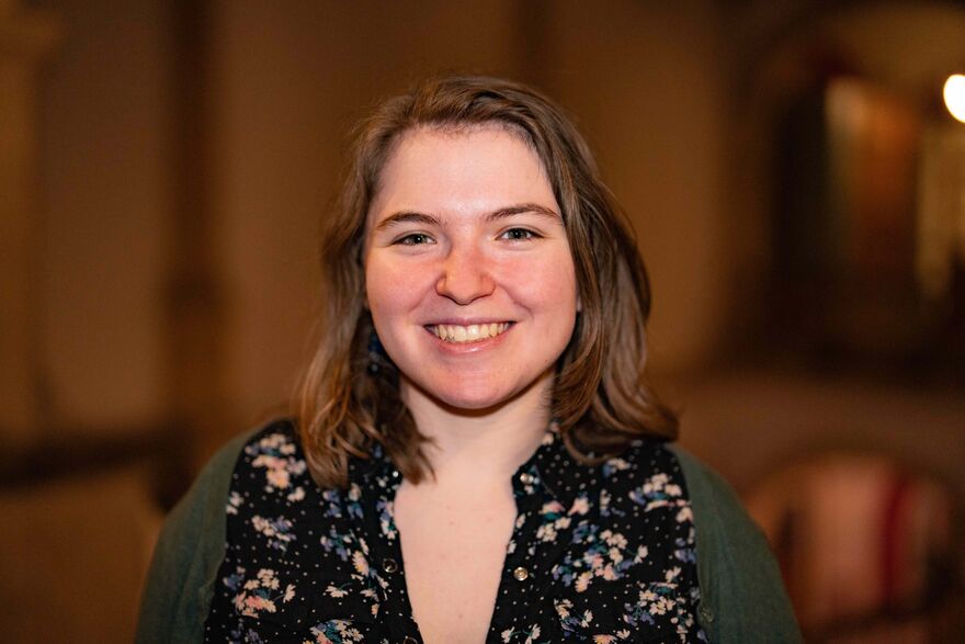 A white woman with a round face and shoulder-length dark blonde hair looks into the camera and smiles, showing her teeth. She is wearing a black collared shirt decorated with a  colourful floral pattern and a green cardigan.