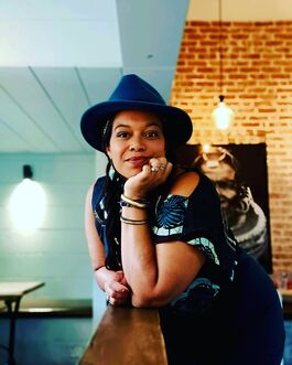 Woman with brown skin, wearing blue hat and blue tshirt, leaning on counter and looking at camera