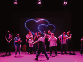 A group of actors on stage watching a man in the centre. The ensemble have shocked expressions on their faces, whilst the man is stood holding a pose with one arm pointing up to the sky. He is wearing a pink feather boa and heart shaped pink sunglasses. The stage has three pink spotlights which cast a neon pink hue over the actors. There are 2 love hearts interlinked in the background.