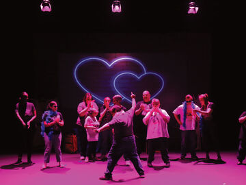 A group of actors on stage watching a man in the centre. The ensemble have shocked expressions on their faces, whilst the man is stood holding a pose with one arm pointing up to the sky. He is wearing a pink feather boa and heart shaped pink sunglasses. The stage has three pink spotlights which cast a neon pink hue over the actors. There are 2 love hearts interlinked in the background.