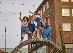 Performers from Autin Dance Theatre standing on a giant wooden wheel wity colourful flag bunting behind