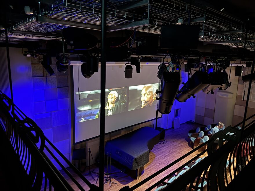 View from a balcony looking down at stage, with theatre spotlights in foreground, an image of Dame Carmen Monroe on screen