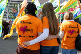 Two people in orange volunteer t-shirts stand facing away from the camera, with their arms around each other. One has long brown hair, the other black. In the background there are colourful flags and another volunteer in a volunteer t-shirt.  