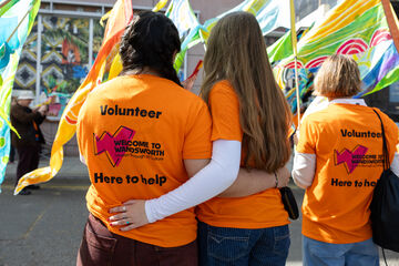 Two people in orange volunteer t-shirts stand facing away from the camera, with their arms around each other. One has long brown hair, the other black. In the background there are colourful flags and another volunteer in a volunteer t-shirt.  