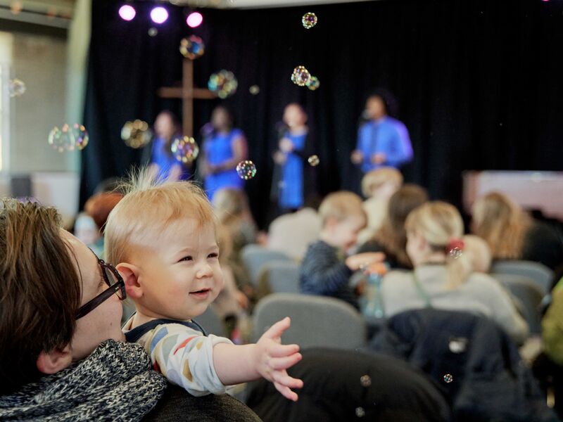 parent holding toddler reaching for bubbles in the air in foreground. Background is other audience members and 4 gospel singers on stage out of focus.
