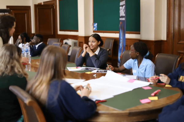 A group of young people sat around a large wooden table working on large sheets of paper