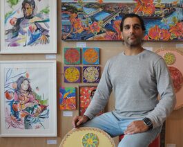 man sitting down in front of paintings on wall with a shield shaped sculpture resting against his leg