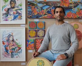 man sitting down in front of paintings on wall with a shield shaped sculpture resting against his leg