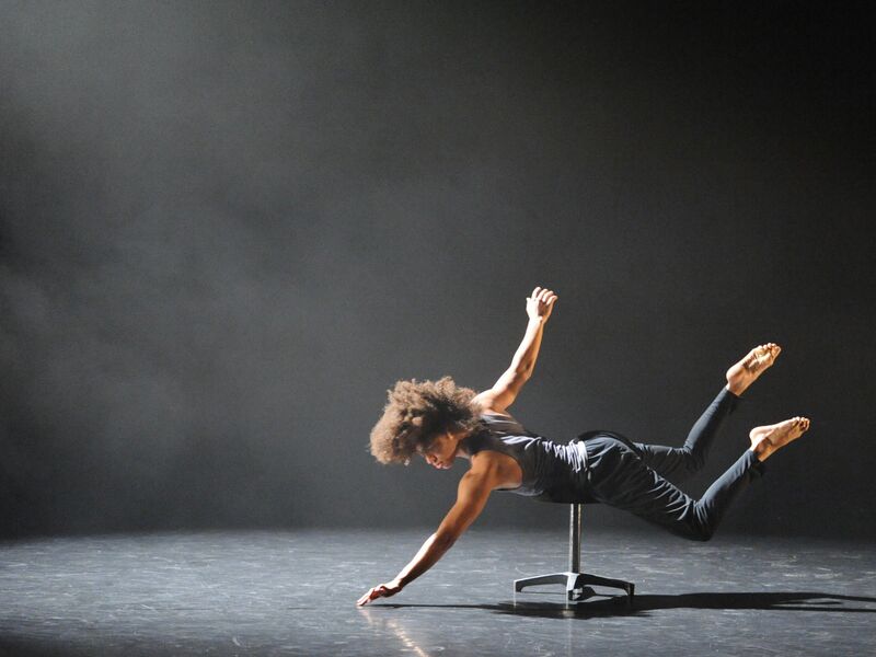 A male dancer lays across a stool while reaching one arm down towards the floor.