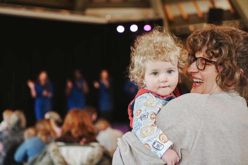 child being held by mother, looking over mothers shoulder at camera. Mother is smiling. Gospel choir performing in background.