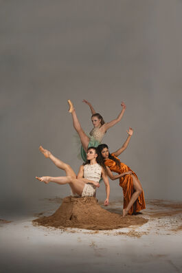 Three women, two are white and one has brown skin, surround a pile of sand against a grey background in a variety of dynamic balletic positions. 