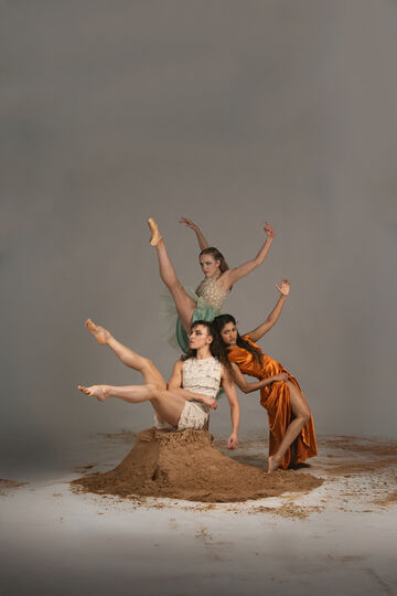 Three women, two are white and one has brown skin, surround a pile of sand against a grey background in a variety of dynamic balletic positions.