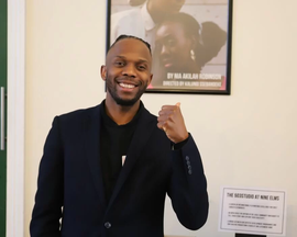 A man posing for a photo in a suit, he is smiling and pointing to the poster on the wall behind him. 