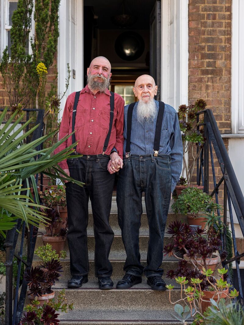 Photographic portrait showing Peter and David holding hands and standing at the foot of stairs. David is in his seventies tall, bald, and has a large nose ring and a beard. Peter is shorter and in his eighties. He is wearing a blue shirt and has a long white beard and is bald.  Both men are white and wearing braces.